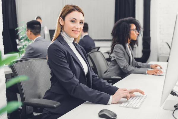 Hermosa joven mujer de negocios utilizando la computadora de escritorio y sonriendo a la cámara en la oficina - foto de stock