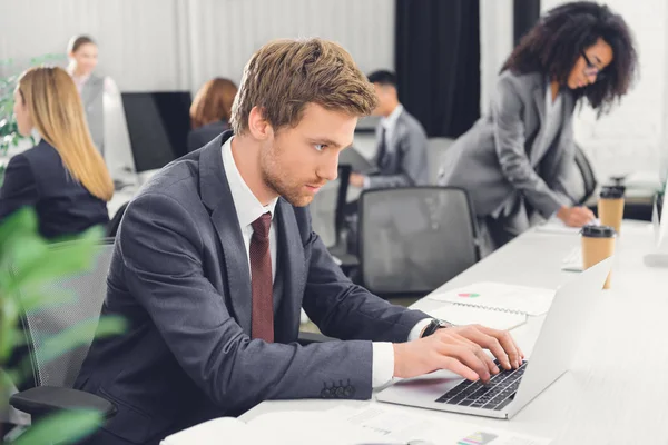 Jeune homme d'affaires concentré utilisant un ordinateur portable tout en travaillant avec des collègues dans un bureau ouvert — Photo de stock