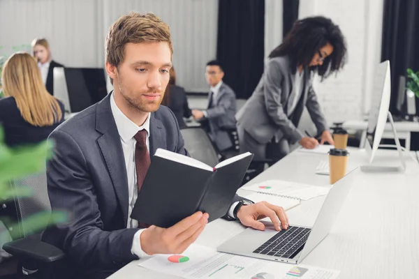 Joven empresario enfocado sosteniendo bloc de notas y utilizando el ordenador portátil en la oficina de espacio abierto - foto de stock