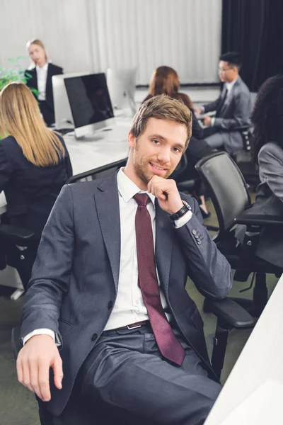 High angle view of handsome young businessman smiling at camera while sitting with hand on chin in open space office — Stock Photo