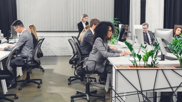 Vista lateral de jóvenes empresarios multiétnicos profesionales que trabajan con dispositivos digitales en oficinas de espacio abierto - foto de stock