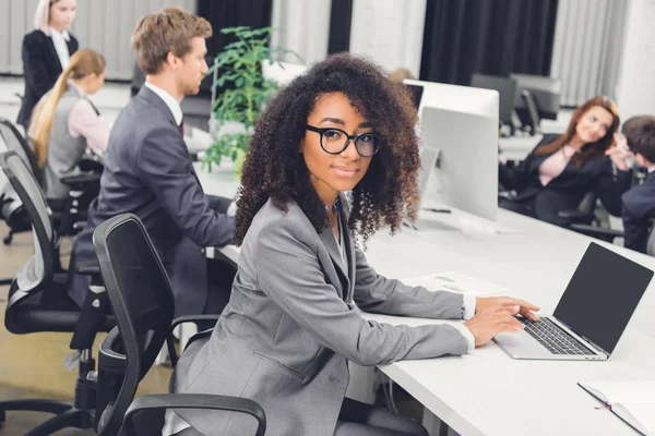 Красива молода афроамериканська бізнес-леді в окулярах посміхається на камеру, використовуючи ноутбук на робочому місці — Stock Photo