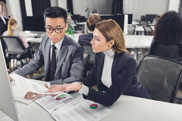 Konzentrierte junge multiethnische Geschäftskollegen, die im Büro mit Papieren und Desktop-Computern arbeiten — Stockfoto