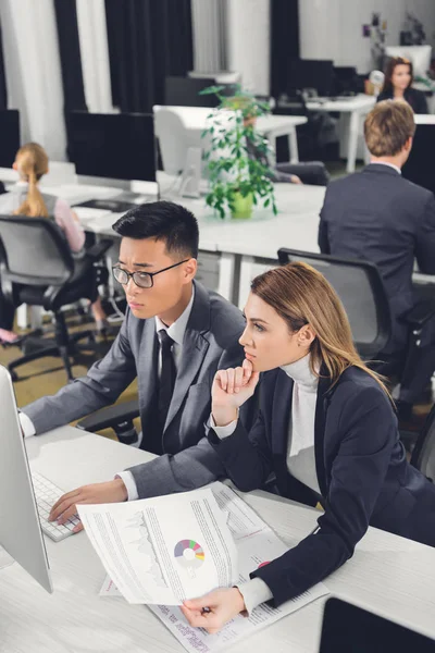 Enfocado joven hombre de negocios y mujer de negocios usando computadora de escritorio y trabajando con papeles en la oficina - foto de stock