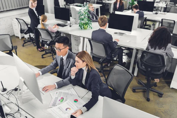 Высокий угол обзора многорасовых предпринимателей, работающих с документами и настольными компьютерами в офисе — стоковое фото
