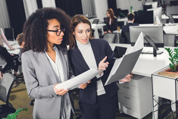 Professionelle junge multiethnische Geschäftsfrauen betrachten Ordner mit Papieren, während sie im Großraumbüro stehen — Stockfoto