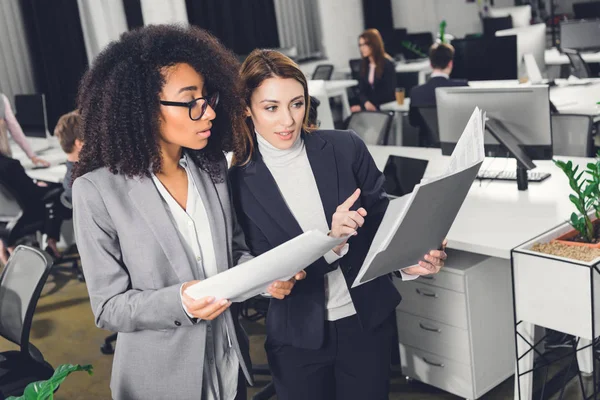 Professionnelles jeunes femmes d'affaires multiethniques regardant le dossier avec des papiers tout en se tenant dans un bureau à aire ouverte — Photo de stock