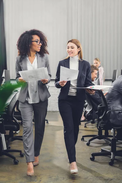 Smiling young multiethnic businesswomen holding papers and walking in office — Stock Photo