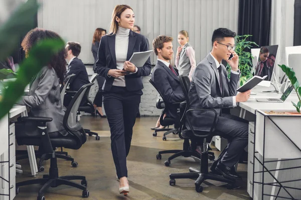 Красива молода бізнес-леді тримає папку з паперами і дивиться на колег під час ходьби в офісі — стокове фото