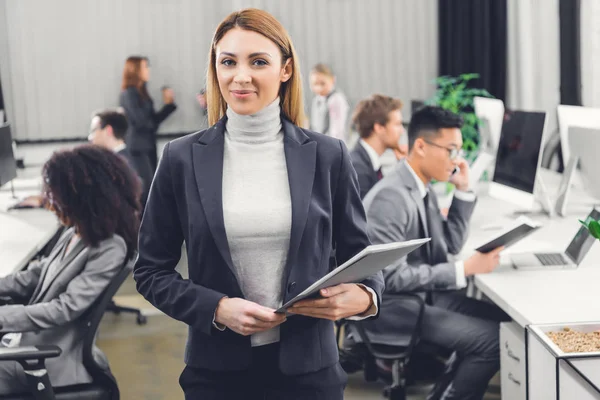 Beautiful young businesswoman holding folder with documents and smiling at camera in office — Stock Photo