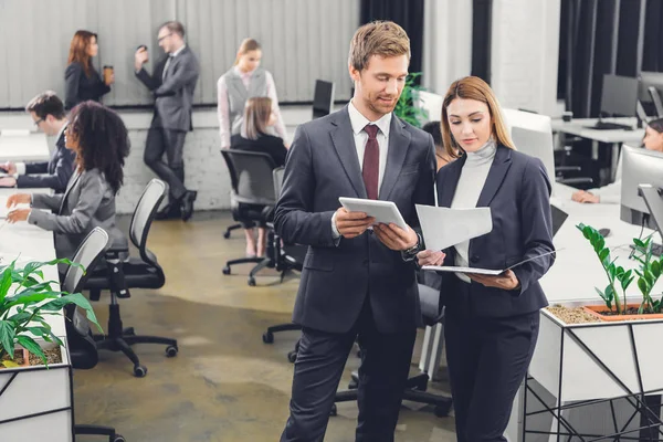 Joven hombre de negocios profesional y mujer de negocios discutiendo papeles mientras están de pie juntos en la oficina de espacio abierto - foto de stock