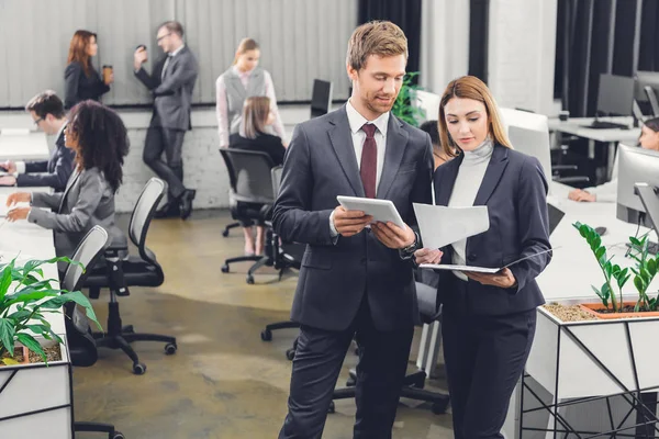 Professionnel jeune homme d'affaires et femme d'affaires discutant des documents tout en se tenant ensemble dans un bureau à aire ouverte — Photo de stock