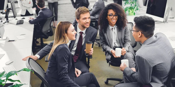 Професійні усміхнені молоді багатоетнічні бізнесмени тримають напої і розмовляють в офісі — стокове фото