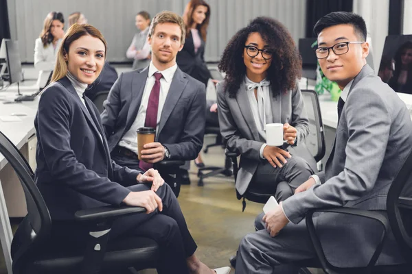 Professionnelle jeune équipe multiraciale d'affaires assis ensemble et souriant à la caméra dans le bureau — Photo de stock