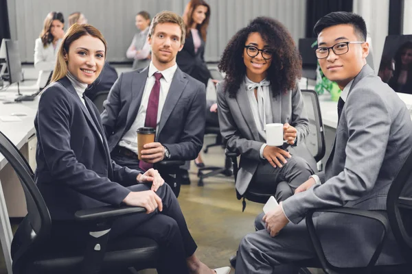 Profesional joven multirracial equipo de negocios sentado juntos y sonriendo a la cámara en la oficina - foto de stock