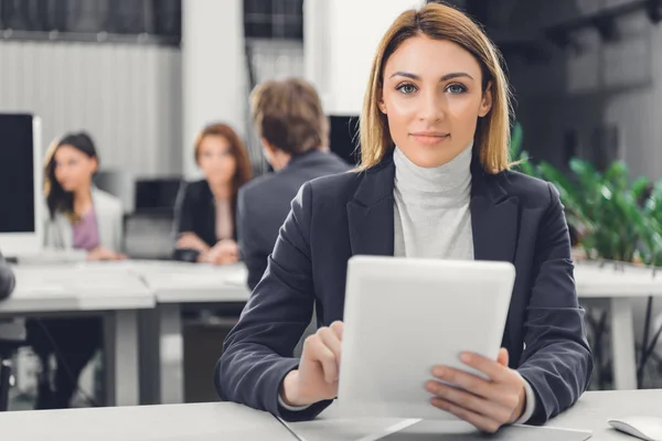 Hermosa joven empresaria sosteniendo tableta digital y sonriendo a la cámara en la oficina - foto de stock