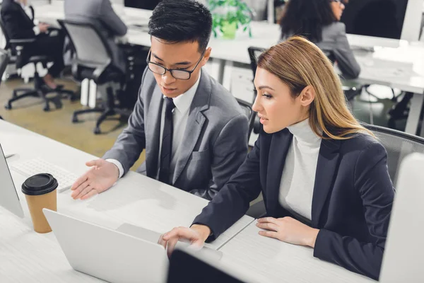 Focused young businesspeople discussing work and pointing at laptop in office — Stock Photo