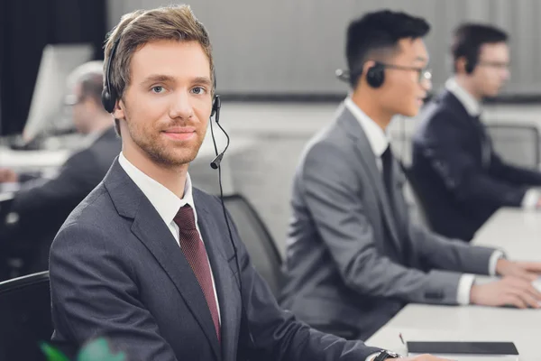 Joven hombre de negocios guapo en auriculares sonriendo a la cámara mientras trabaja con colegas en la oficina - foto de stock