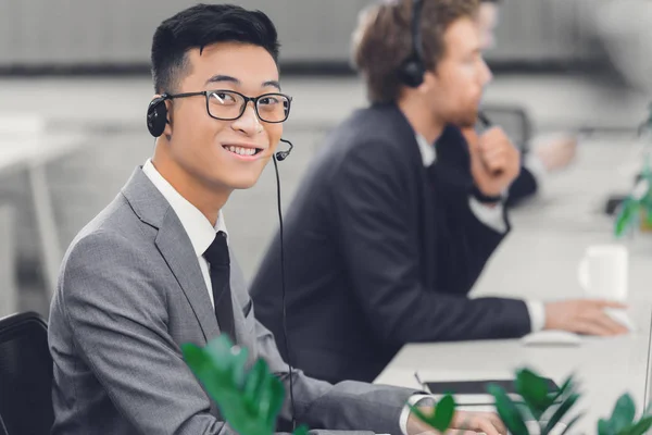 Guapo joven asiático hombre de negocios en auriculares sonriendo a cámara mientras trabaja en call center - foto de stock