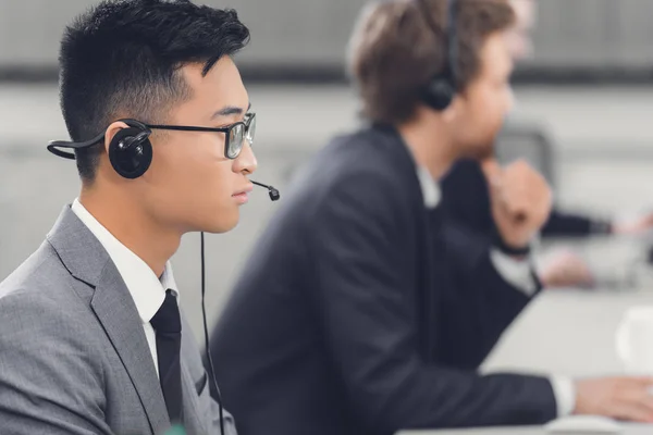 Vista laterale di focalizzato giovane uomo d'affari asiatico in cuffia lavorando con i colleghi in ufficio — Foto stock