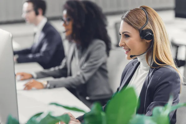 Visão lateral de sorrir jovem empresária em fone de ouvido trabalhando com colegas em call center — Fotografia de Stock