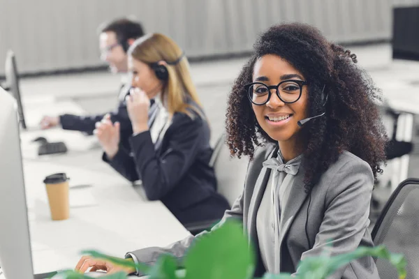 Jovem mulher americana africana atraente em fone de ouvido sorrindo para a câmera enquanto trabalhava com colegas no escritório — Fotografia de Stock