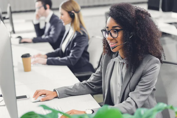 Jeune femme d'affaires afro-américaine souriante dans un casque de travail avec des collègues au bureau — Photo de stock