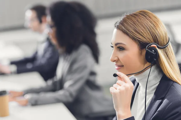 Side view of smiling young woman in headset working with colleagues in office — Stock Photo