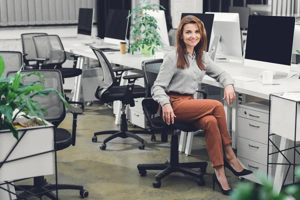 Atraente jovem empresária sentado no local de trabalho e sorrindo para a câmera — Fotografia de Stock