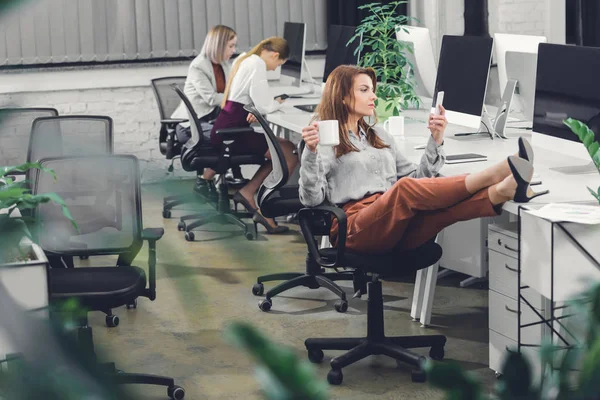 Hermosa joven empresaria sosteniendo la taza y el uso de teléfono inteligente mientras está sentado con las piernas en el escritorio en la oficina de espacio abierto - foto de stock