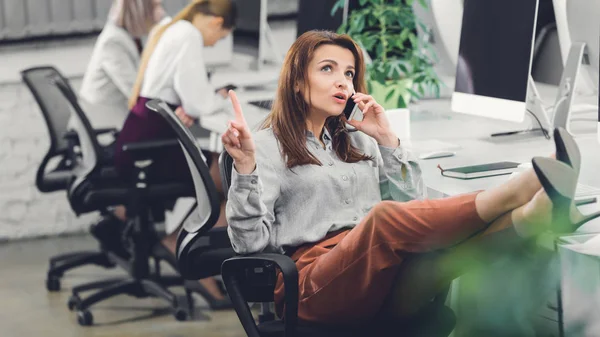 Bela jovem empresária apontando para cima com o dedo e falando por smartphone no escritório — Fotografia de Stock