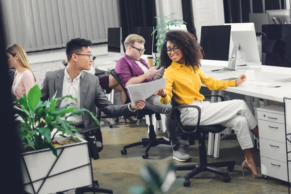 Jóvenes empresarios multirraciales sonrientes sosteniendo papeles y trabajando juntos en la oficina - foto de stock