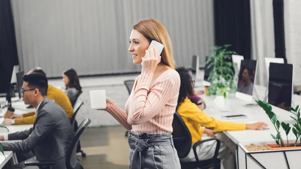 Усміхнена молода бізнес-леді тримає чашку і розмовляє по смартфону, поки колеги працюють в офісі — стокове фото