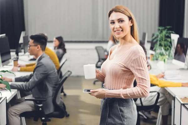 Hermosa joven mujer de negocios con taza y teléfono inteligente sonriendo a la cámara mientras los colegas que trabajan detrás en la oficina - foto de stock