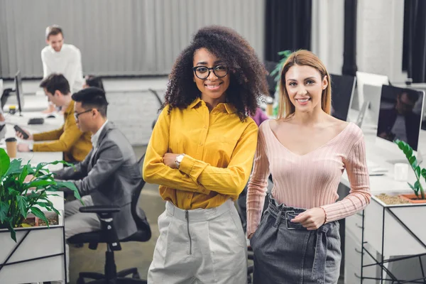 Hermosas empresarias multiétnicas de pie y sonriendo a la cámara mientras sus colegas trabajan detrás en la oficina - foto de stock