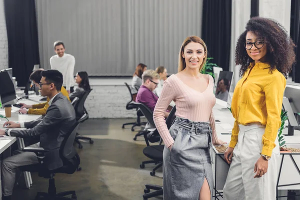 Jóvenes empresarias multiétnicas sonriendo a la cámara mientras sus colegas trabajan detrás en la oficina - foto de stock