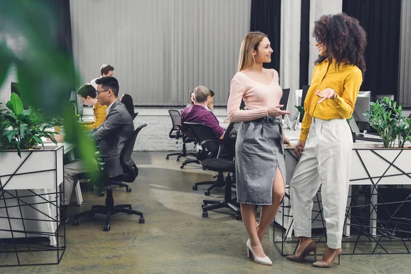 Giovani imprenditrici multietniche che parlano e si sorridono a vicenda mentre i colleghi lavorano dietro in ufficio — Foto stock