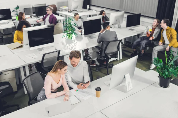 Visão de alto ângulo do jovem empresário e empresária usando smartphone enquanto colegas sentados e trabalhando no escritório — Fotografia de Stock