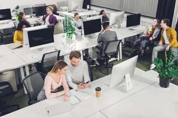 Blick aus der Vogelperspektive auf junge Geschäftsfrau und Geschäftsfrau mit Smartphone, während Kollegen im Büro sitzen und arbeiten — Stockfoto