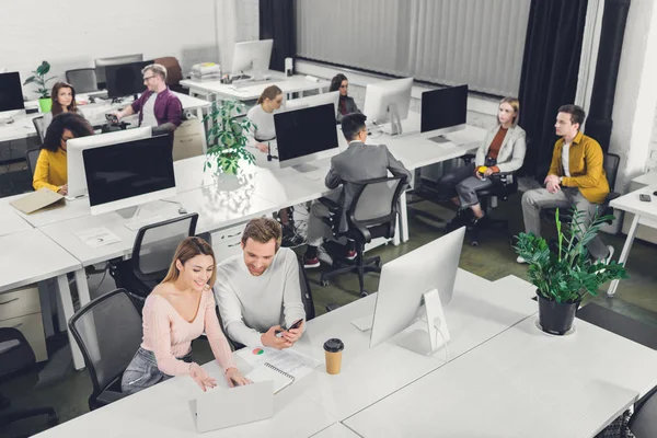 Vista de ángulo alto de los jóvenes colegas de negocios sentados y trabajando juntos en la oficina de espacio abierto - foto de stock