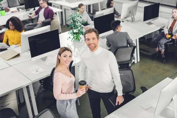 Vue grand angle de jeunes gens d'affaires joyeux souriant à la caméra tout en se tenant debout dans un bureau à aire ouverte — Photo de stock