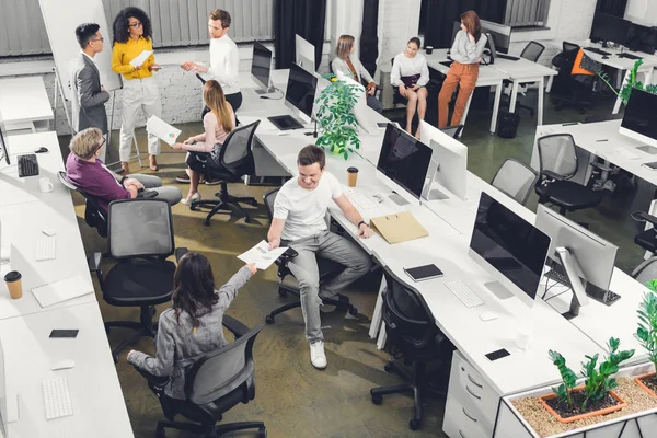 Vista de ángulo alto de colegas jóvenes profesionales que trabajan con papeles y computadoras en oficinas de espacio abierto - foto de stock