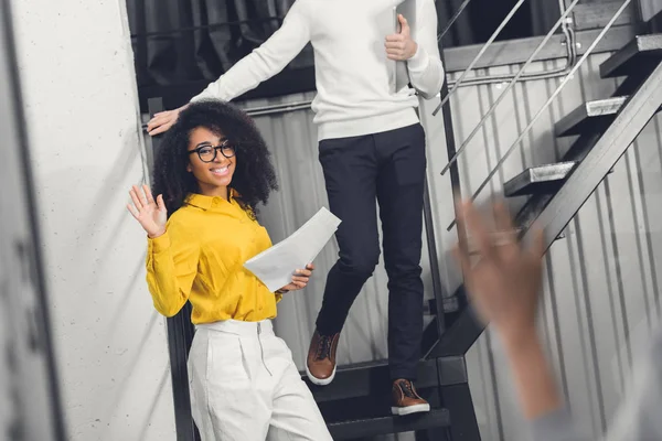 Cropped shot of smiling african american businesswoman holding papers and waving hand to colleague in office — Stock Photo