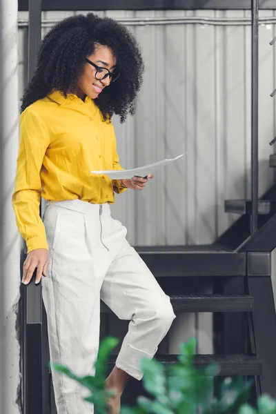 Fröhliche junge afrikanisch-amerikanische Geschäftsfrau hält Papiere in der Hand und steht im Büro auf einer Treppe — Stockfoto