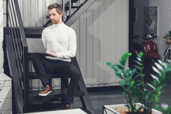 Konzentrierter junger Mann sitzt auf Treppe und benutzt Laptop im Büro — Stockfoto
