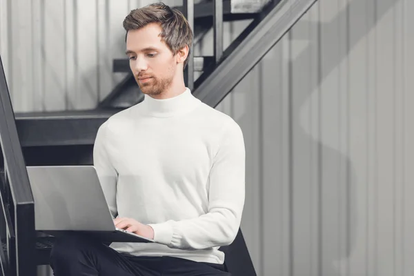 Зосереджений молодий бізнесмен сидить на сходах і використовує ноутбук в офісі — стокове фото
