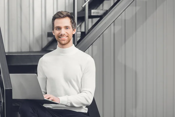 Handsome young businessman using laptop and smiling at camera in office — Stock Photo