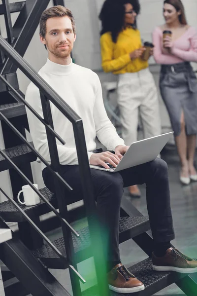 Красивий чоловік сидить з ноутбуком на сходах і посміхається на камеру, а жінки-колеги п'ють каву позаду в офісі — стокове фото