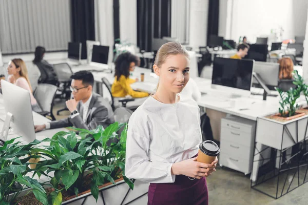 Beautiful young businesswoman holding coffee to go and smiling at camera while colleagues working behind in open space office — Stock Photo