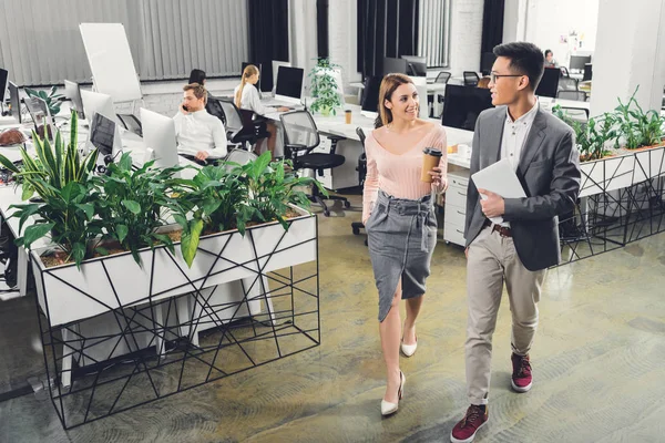 Vista de ángulo alto de los jóvenes empresarios con tableta digital y café para ir caminando y sonriendo unos a otros en la oficina - foto de stock